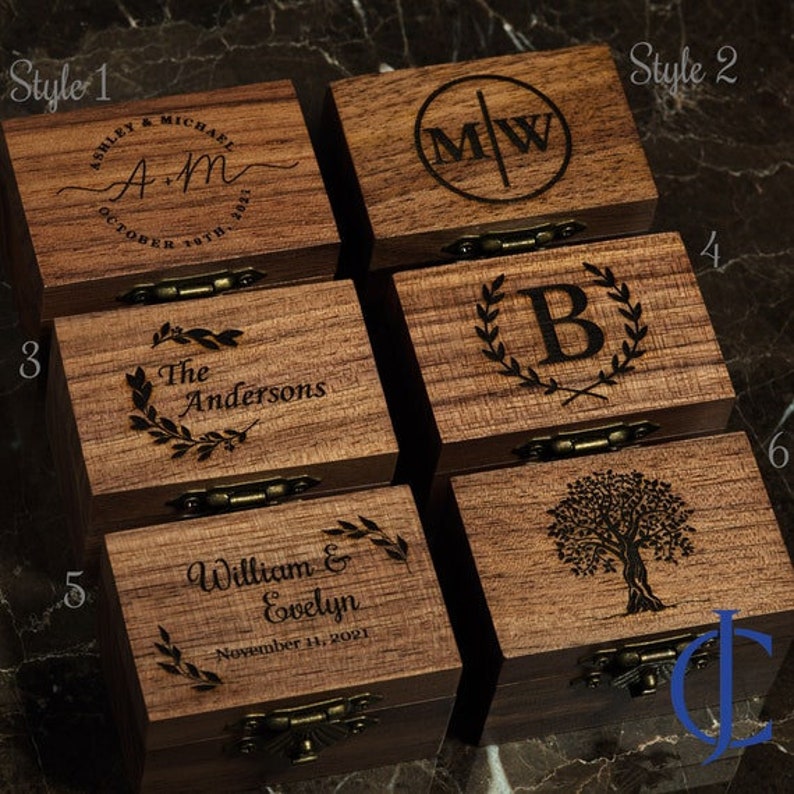 May include: Six wooden ring boxes with different engraved designs. The boxes are made of dark wood and have a hinged lid. The designs include a monogram, a tree, a wreath, and a couple's names and wedding date.