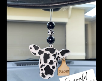 May include: Black and white cow head car charm with a wooden tag that says "Kacey". The charm is hanging from a rearview mirror with black and white beads.