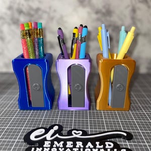 May include: Three pencil-sharpener-shaped pen holders in blue, purple, and gold, each filled with pens and pencils. The blue holder contains glitter pencils, the purple has pens, and the gold has pens. A black and white logo for "Emerald Innovations, LLC" is at the bottom.