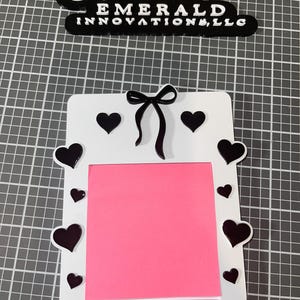 May include: White frame with a pink sticky note inside. The frame is decorated with black hearts and a black bow. The text "EMERALD INNOVATIONS, LLC" is visible at the top of the image.
