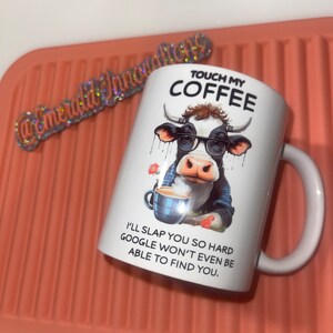 May include: White ceramic coffee mug with a black and white cow wearing sunglasses and holding a cup of coffee. The mug has the text "Touch My Coffee" and "I'll slap you so hard Google won't even be able to find you."