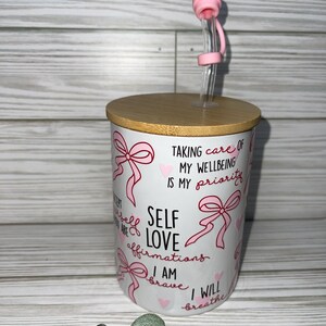 May include: White ceramic mug with a pink and red design. The mug has a bamboo lid and a pink silicone straw. The mug has the text "Self Love Affirmations" and "I am brave I will breathe" printed on it.