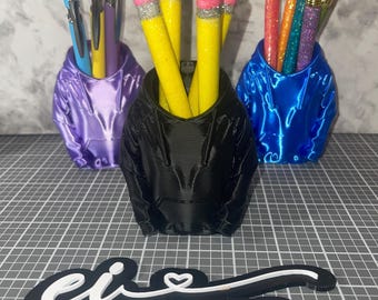 May include: Three pen holders shaped like hoodies, in purple, black, and blue, each filled with pens and pencils. The pencils are yellow with pink erasers and silver glitter. The pens are various colors. The logo for Emerald Innovations, LLC is at the bottom.