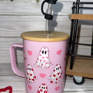 May include: Pink mug with a handle, featuring a pattern of white ghosts decorated with red hearts and flowers. The mug has a wooden lid and a clear straw with a black stopper. A Valentine's Day themed drinkware.