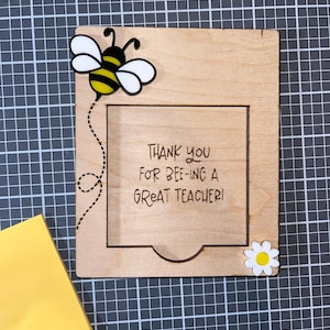 May include: Wooden frame with a bee and flower design. The frame has a cutout for a photo or message. The text "Thank you for bee-ing a great teacher!" is engraved on the frame.