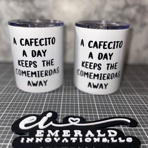 May include: Two white insulated tumblers with black text that reads "A Cafecito A Day Keeps The Comemierdas Away". The tumblers are sitting on a black and white checkered surface. The text is in a bold, sans-serif font.