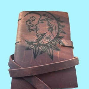 May include: A brown leather journal with a wrap-around strap. The cover features a black illustration of a woman's profile within a crescent moon, smoking. The design includes leaves and circular patterns.