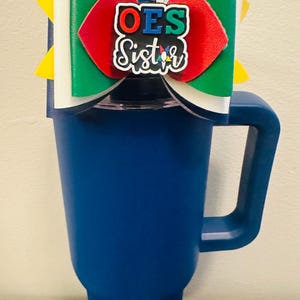 Puede incluir: Un vaso azul con asa, coronado por un lazo decorativo. El lazo presenta secciones verdes, rojas y blancas, con el texto "OES Sister" en el centro. Se ven detalles amarillos en los lados del lazo.