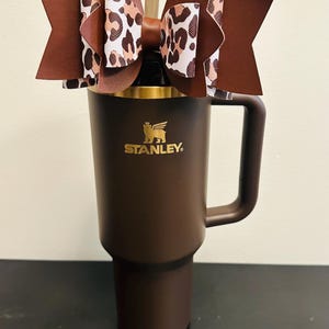 Puede incluir: Un vaso Stanley marrón con asa, coronado con un lazo decorativo. El lazo presenta un diseño marrón y estampado de leopardo. El vaso tiene el logotipo de Stanley en dorado.