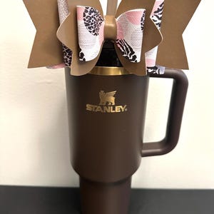 Puede incluir: Un vaso Stanley marrón con un lazo decorativo. El lazo presenta tela estampada marrón, rosa y blanca. El vaso tiene un asa y una pajita. El logotipo de Stanley es visible en dorado.