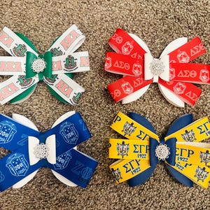 Puede incluir: Cuatro lazos decorativos para el cabello en verde, rojo, azul y amarillo. Cada lazo presenta un adorno central y cintas impresas con letras y símbolos griegos. Los lazos están dispuestos sobre una superficie texturizada.