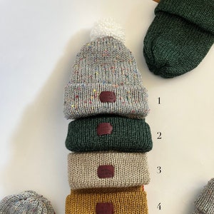 May include: Five knitted beanies in different colors, including grey, green, beige, mustard yellow, and rust orange. Each beanie has a small leather patch with the text "Knit With Love" on it. The top beanie has a white pompom.