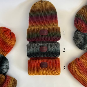 May include: Three knitted hats in different color combinations of orange, red, green, and black. The hats are stacked on top of each other. Each hat has a small leather tag with the text "Knit &  Co."