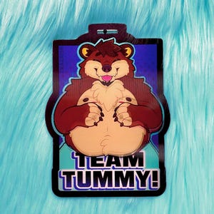May include: A rectangular badge featuring a cartoon bear with a brown body, tan belly, and a smiling expression. The badge has the text "TEAM TUMMY!" in white and purple. The background is blue.