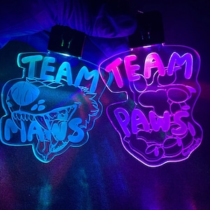 May include: Two illuminated acrylic signs with the words "TEAM MAWS" and "TEAM PAWS" in a stylized font. The "MAWS" sign features a cartoon monster face, while the "PAWS" sign has a paw print design. Both signs glow with vibrant colors.