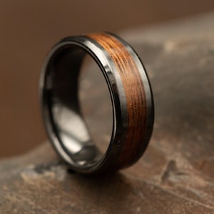May include: A close-up of a black ring with a wooden inlay. The ring has a polished black band with a silver-coloured edge. The wooden inlay features a warm brown colour with visible wood grain.