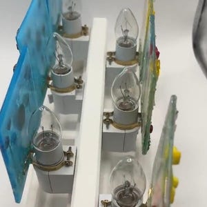 May include: A decorative light fixture with six clear light bulbs. The fixture has a white base and features colorful glass panels with various designs. The glass panels are blue, green, and yellow.