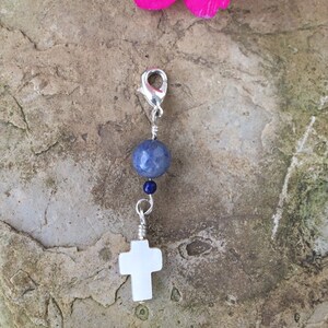 May include: A silver charm with a white cross and a blue gemstone bead. The charm is hanging from a silver clasp. The charm is on a stone background with a pink flower.