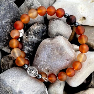 Könnte beinhalten: Ein Armband mit orangefarbenen und bernsteinfarbenen Perlen, akzentuiert mit dunkelgrauen Perlen und einem silbernen Anhänger. Das Armband liegt auf einem Bett aus glatten, grauen und weißen Steinen.