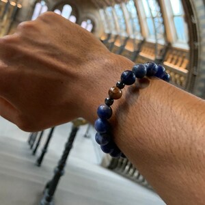 May include: A blue and brown beaded bracelet with a dark blue background. The bracelet is made of round beads and is worn on a person's wrist.