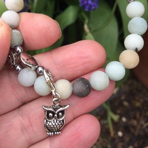 May include: A bracelet made of light green, brown, and white beads with a silver owl charm. The bracelet is on a hand with a blurry background of purple flowers.