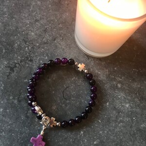 May include: A purple and black beaded bracelet with a silver cross charm. The bracelet is on a gray surface next to a lit candle.