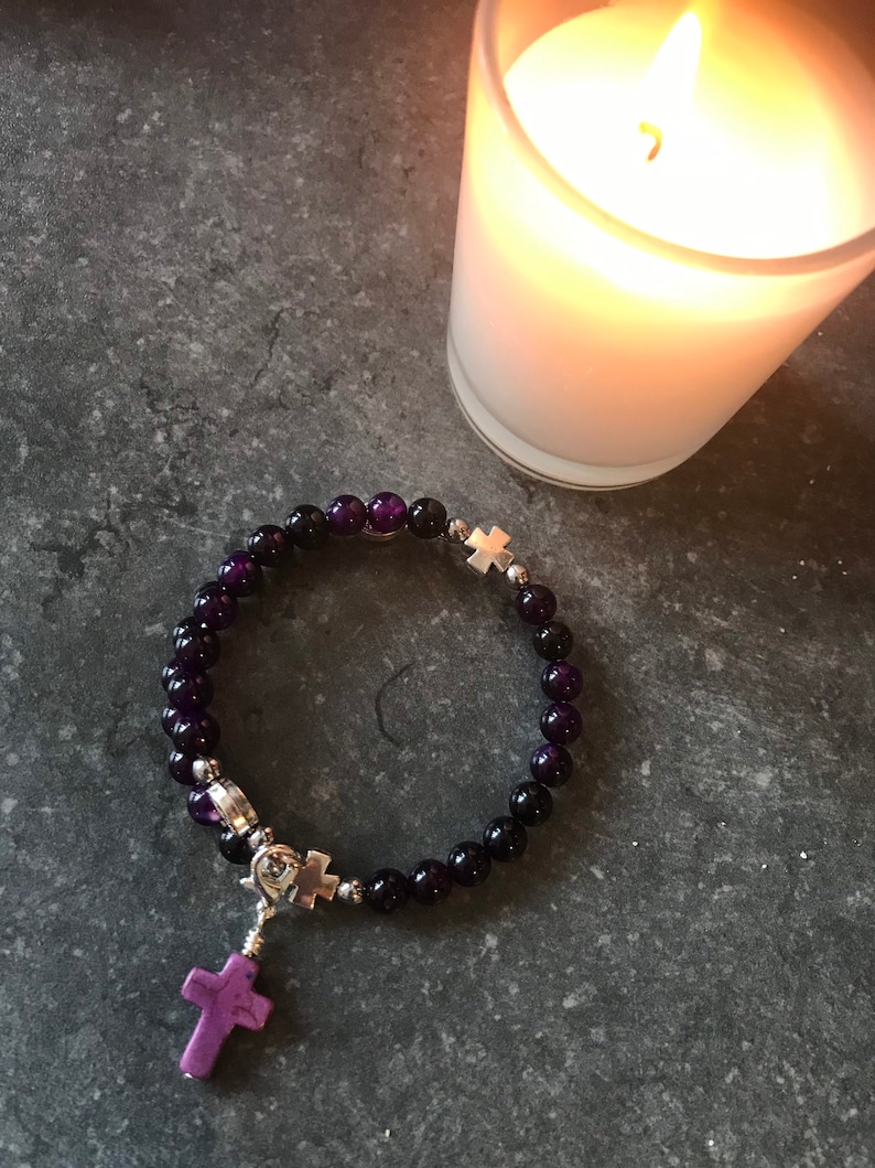 May include: A purple and black beaded bracelet with a silver cross charm. The bracelet is on a gray surface next to a lit candle.