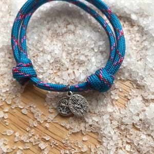 Könnte beinhalten: Ein blaues und pinkfarbenes geflochtenes Armband aus Seil mit einem silbernen Anhänger mit einem Kreuz und einem religiösen Symbol. Das Armband liegt auf einem weißen Hintergrund.