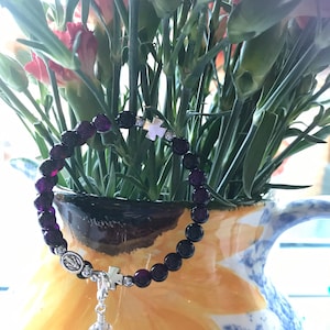 May include: A purple beaded bracelet with a silver charm featuring a cross and a religious icon. The bracelet is lying on a yellow and blue floral patterned pitcher.