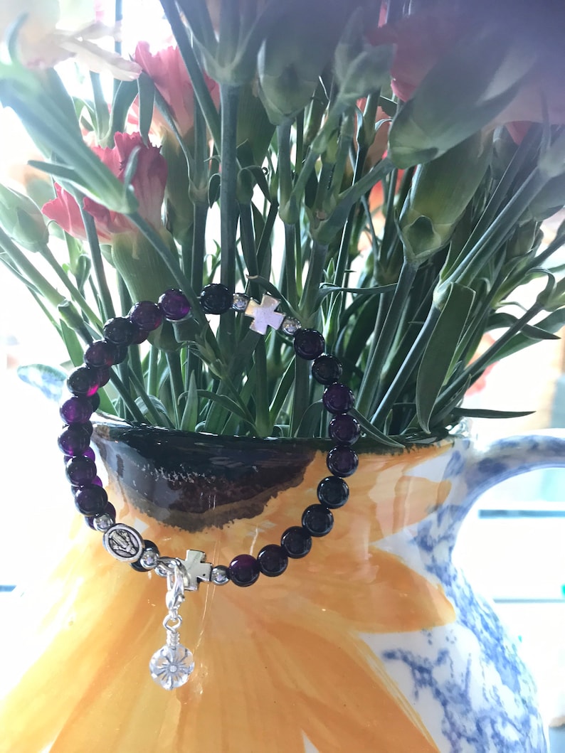 May include: A purple beaded bracelet with a silver charm featuring a cross and a religious icon. The bracelet is lying on a yellow and blue floral patterned pitcher.