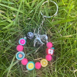 May include: A colorful fruit-themed keychain with a silver key ring. The keychain features a smiley face, a bird, a slice of grapefruit, a watermelon, a lemon, and an apple.