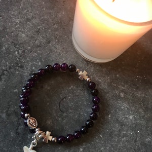May include: A purple amethyst beaded bracelet with silver charms, including a cross, a butterfly, and a depiction of the Virgin Mary. The bracelet is on a gray surface next to a lit candle.