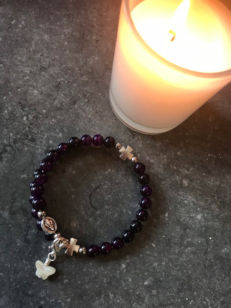May include: A purple amethyst beaded bracelet with silver charms, including a cross, a butterfly, and a depiction of the Virgin Mary. The bracelet is on a gray surface next to a lit candle.