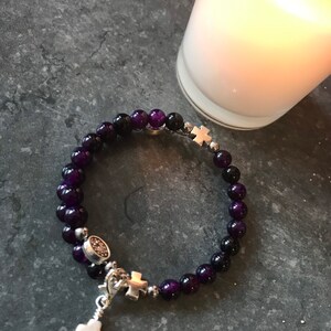 May include: A purple and black beaded bracelet with silver cross charms. The bracelet has a small silver charm with a cross and a larger silver cross charm hanging from it.