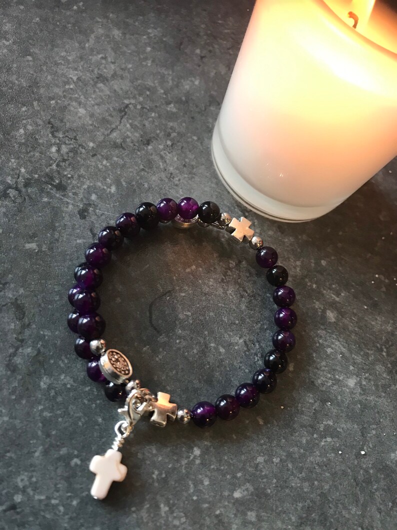 May include: A purple and black beaded bracelet with silver cross charms. The bracelet has a small silver charm with a cross and a larger silver cross charm hanging from it.