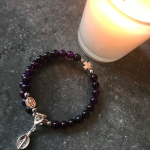 May include: A purple amethyst beaded bracelet with a silver charm of the Virgin Mary and a silver cross charm.