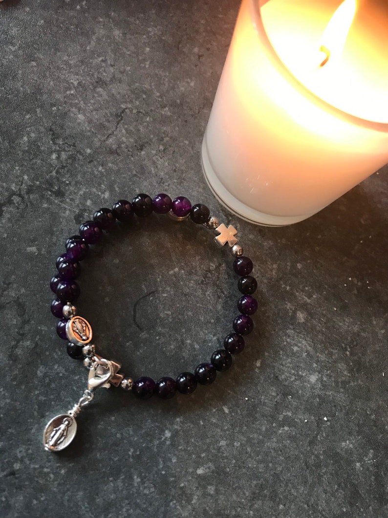 May include: A purple amethyst beaded bracelet with a silver charm of the Virgin Mary and a silver cross charm.