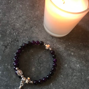May include: A purple gemstone bracelet with silver cross charms and a silver pendant featuring a religious icon. The bracelet is on a gray surface next to a white candle.
