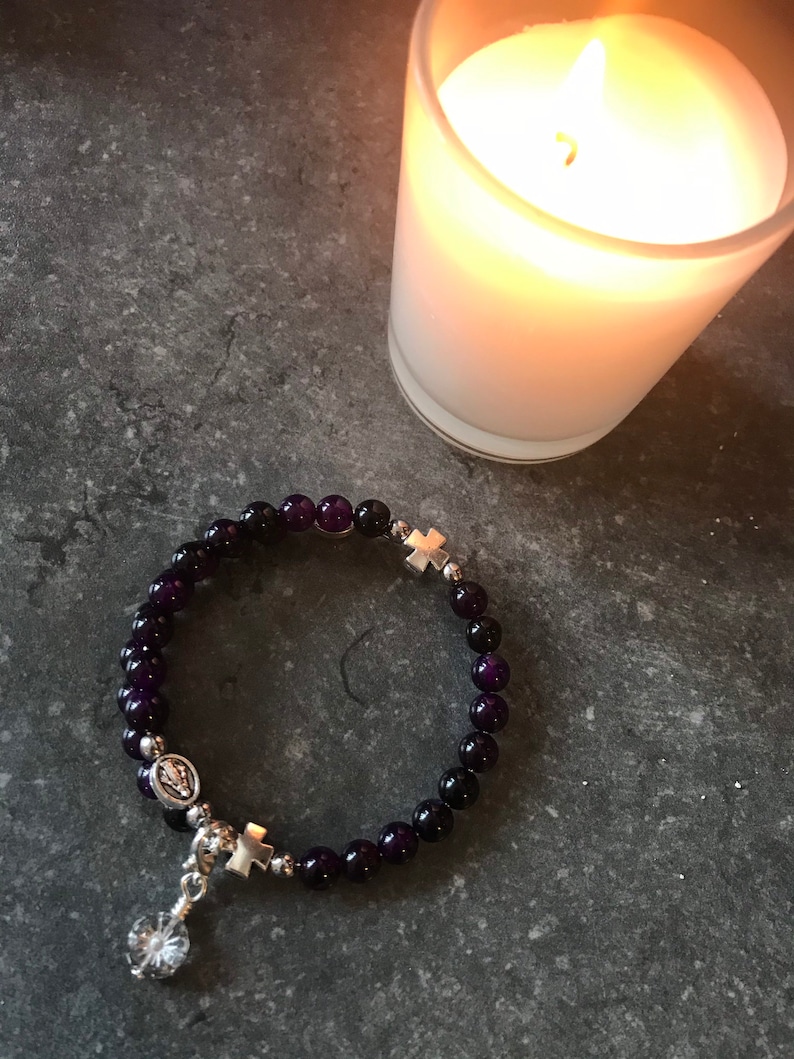 May include: A purple gemstone bracelet with silver cross charms and a silver pendant featuring a religious icon. The bracelet is on a gray surface next to a white candle.
