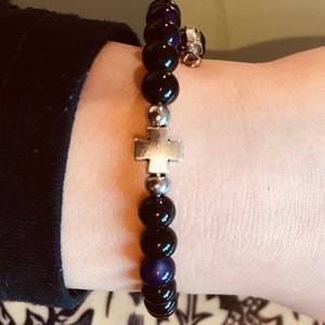 May include: A black and purple beaded bracelet with a silver cross charm. The bracelet is on a person's wrist.