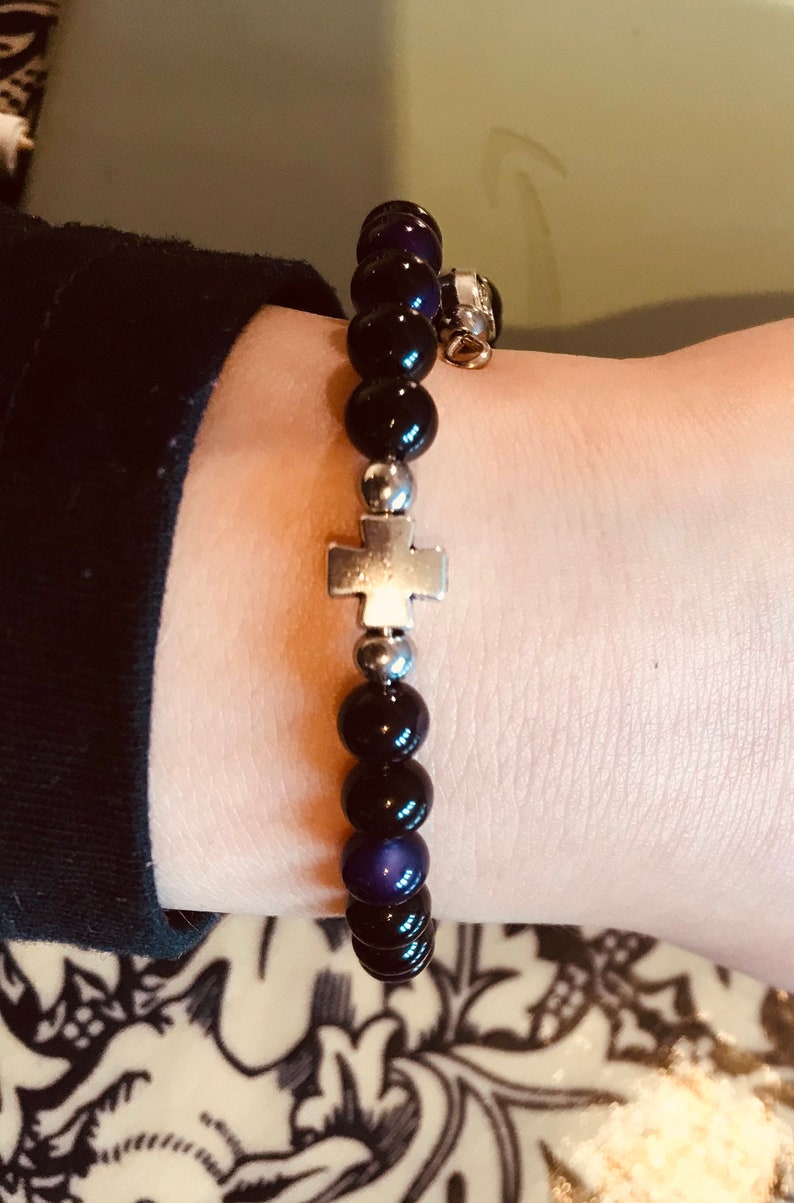 May include: A black and purple beaded bracelet with a silver cross charm. The bracelet is on a person's wrist.