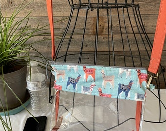 May include: A clear plastic crossbody bag with a zipper closure. The bag has a red and white patterned fabric lining with a llama design. The bag is being displayed on a black wire mannequin.