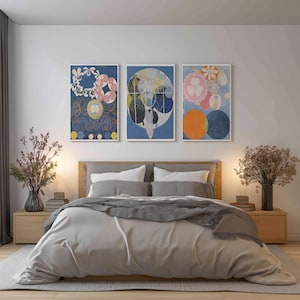 May include: Three framed abstract art prints with circular and floral designs in blue, pink, and yellow hues hang above a bed. The bed has a light gray comforter, pillows, and a wooden headboard. Nightstands with lamps and vases flank the bed.