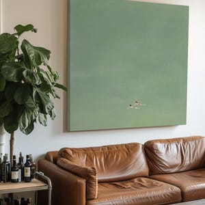 Op de afbeelding: Een groot vierkant canvas kunstwerk met twee figuren die zwemmen in een groene watermassa. Het kunstwerk hangt boven een bruine leren bank. Een potplant en een barkar met flessen staan op de voorgrond.