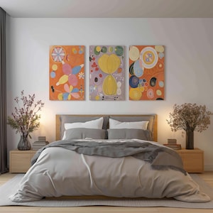 May include: A bedroom scene featuring a bed with a gray blanket and pillows, flanked by wooden nightstands. Above the bed, three abstract art pieces with orange, yellow, and blue hues hang on the wall. The room is decorated with vases of dried flowers.