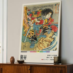 Op de afbeelding: Een ingelijste Japanse houtsnede met een krijger in kleurrijke kleding, die een zwaard vasthoudt. Het kunstwerk heeft Japanse tekst en wordt tentoongesteld op een houten kast. De prent maakt deel uit van de serie "Verhalen van Dappere Krijgers."