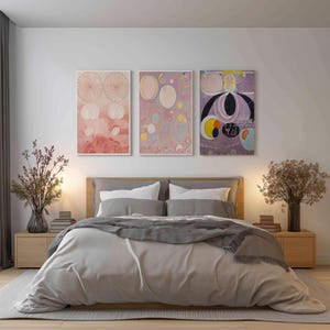 May include: A bedroom scene featuring a bed with a light gray comforter and pillows. Above the bed are three framed abstract art pieces in pink, purple, and yellow hues. Wooden nightstands flank the bed, each with a lamp and decorative vase.