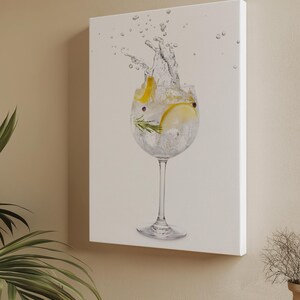 May include: A canvas print showing a cocktail with a splash. A large glass contains ice, lemon slices, and herbs, with water spraying upwards. The background is off-white, and the print is mounted on a wooden frame.