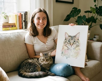 May include: A watercolor painting of a cat with green eyes and a fluffy coat is held by a person. A live cat with similar coloring sits on the couch next to the person. The painting has soft pink and green accents.