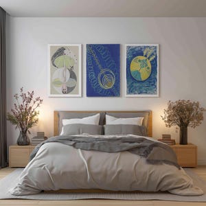 May include: A bedroom scene featuring a bed with a light gray comforter and pillows. Above the bed are three framed abstract art prints in white frames. The room is decorated with wooden nightstands, lamps, and vases with dried flowers.